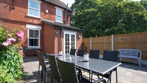 A patio with a table and chairs, a brick house, a wooden fence, and a bench. - Finest Retreats | House on The Green (Derby)