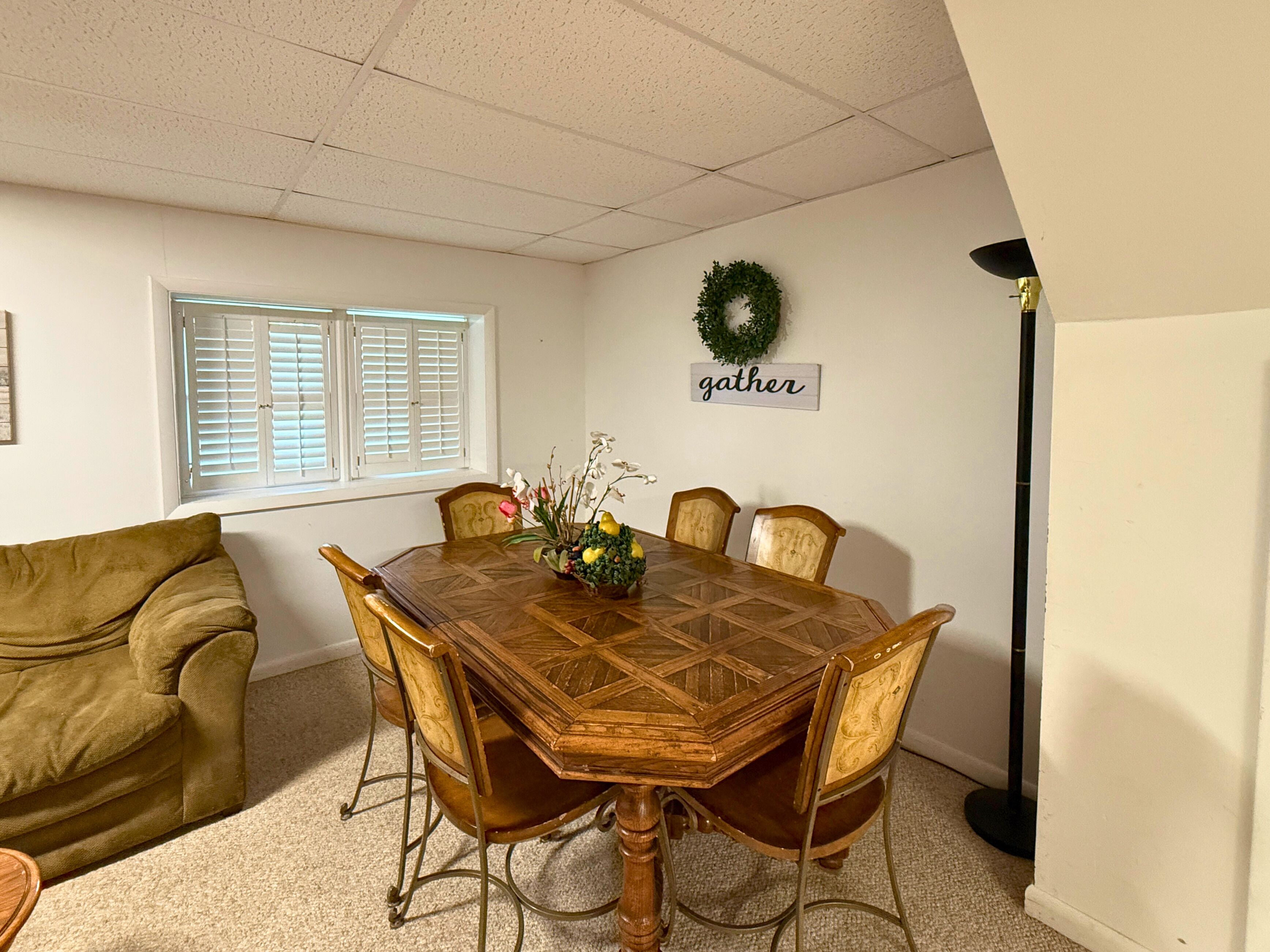 A dining area with a wooden table, six chairs, a green sofa, a window with blinds, a wall-mounted wreath, and a 'gather' sign.