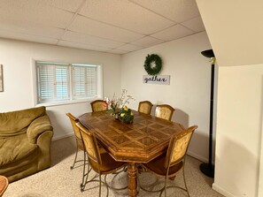Un comedor con una mesa de madera, seis sillas, un sofá verde, una ventana con persianas, una guirnalda en la pared y un letrero que dice 'gather'.