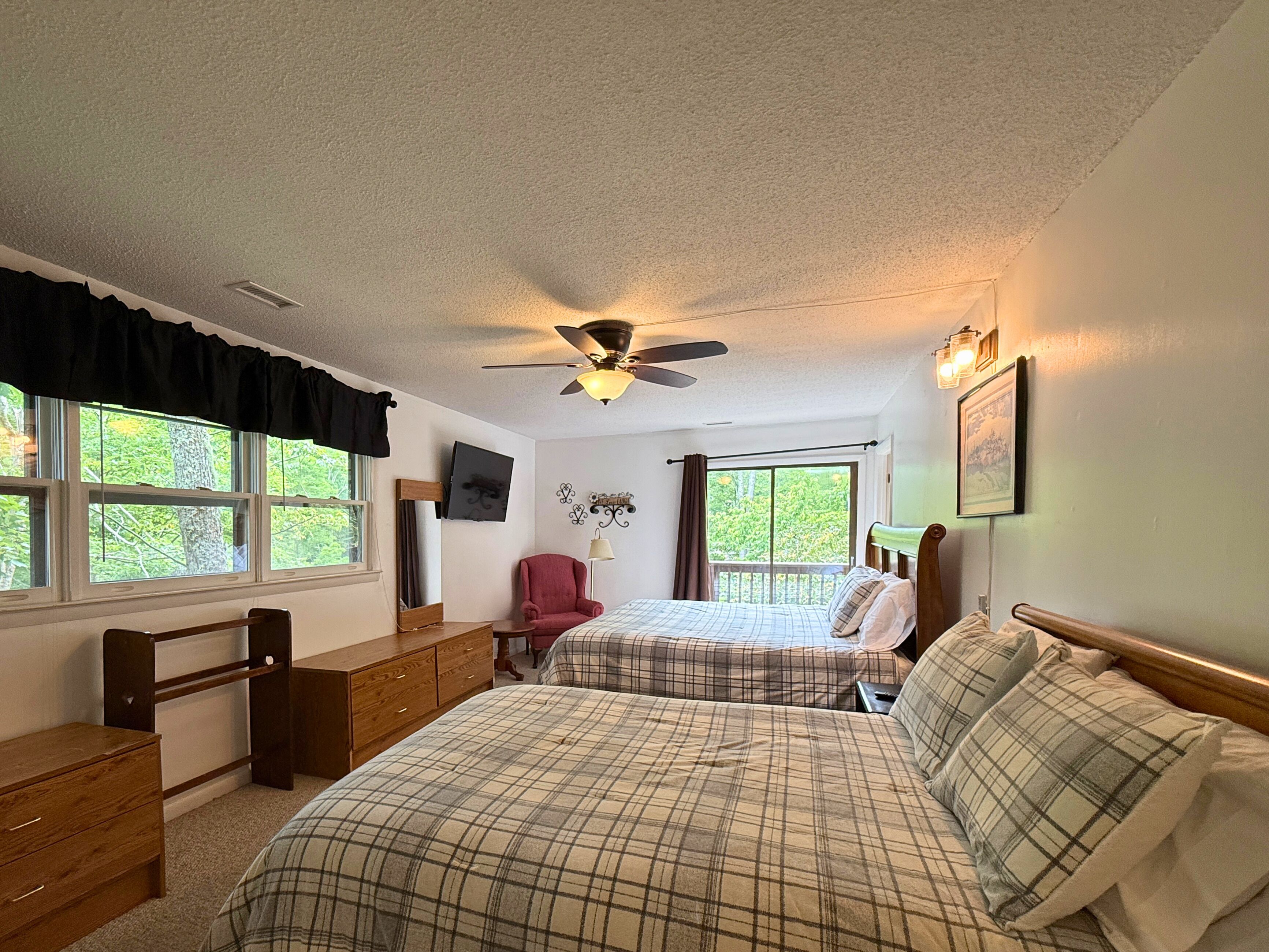 A bedroom with two beds, a ceiling fan, a dresser, a chair, and a window with a view of trees.