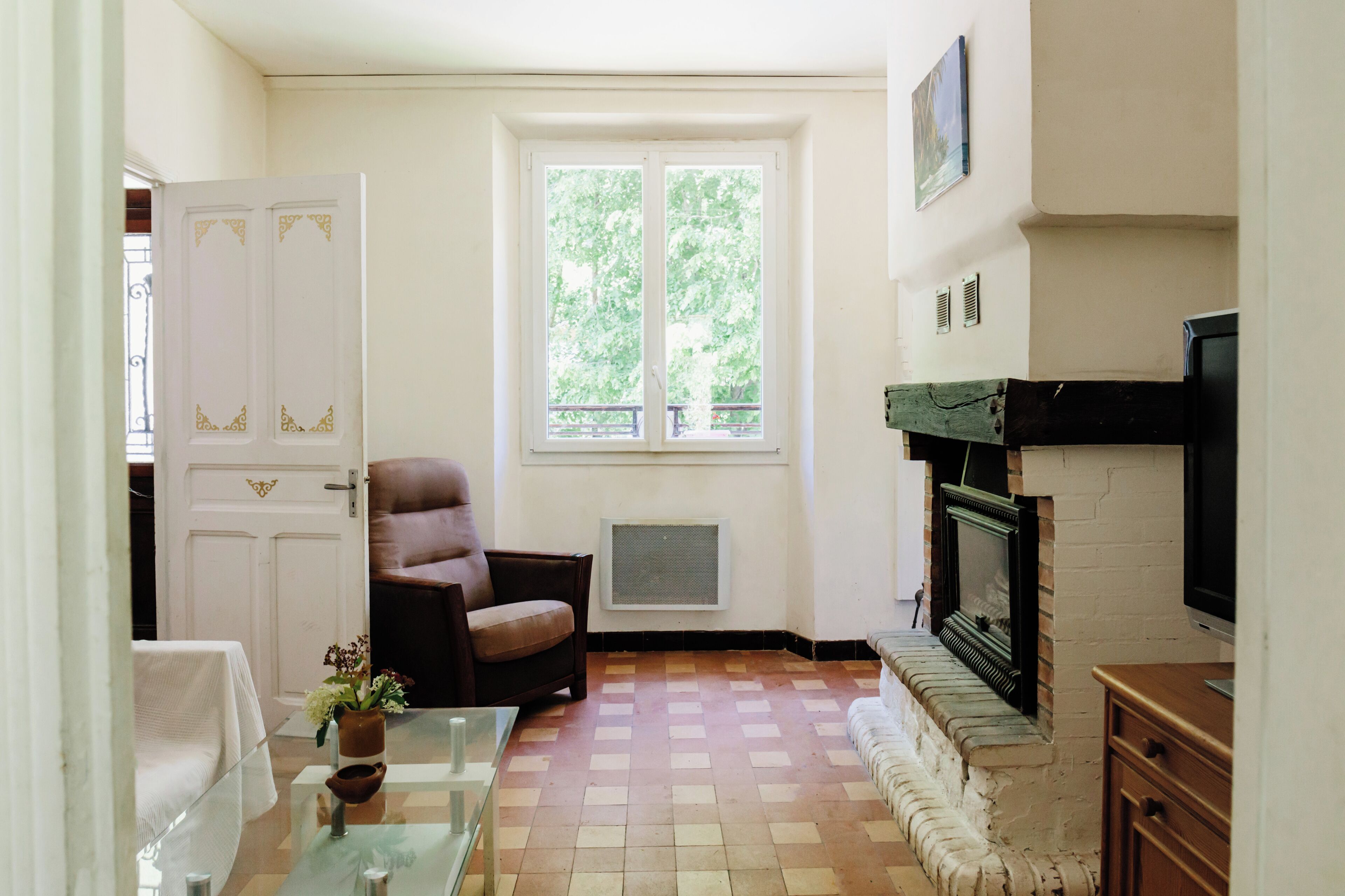 Une pièce avec une cheminée, une fenêtre donnant sur un paysage verdoyant, une porte agrémentée d’éléments décoratifs et un fauteuil.