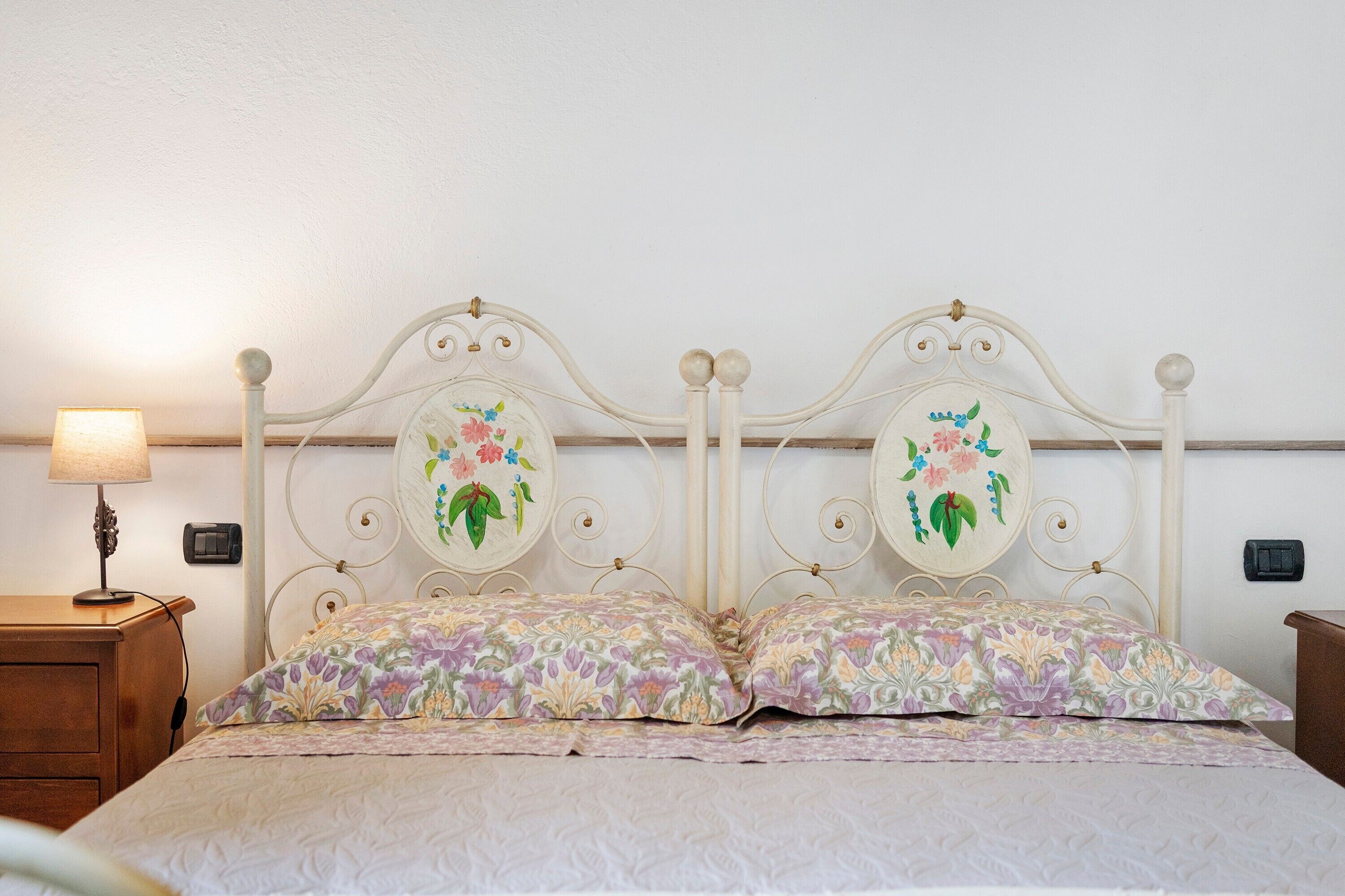 Un lit double avec une tête de lit à motifs floraux et des coussins assortis, une table de chevet avec une lampe et un interrupteur fixé au mur.