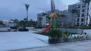 Ein Spielplatz mit einer roten Rutsche, Palmen und modernen Gebäuden im Hintergrund.