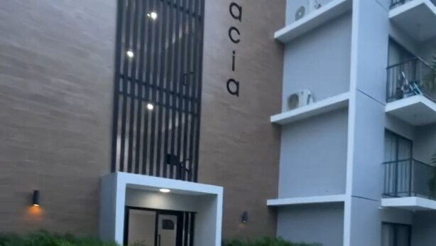 Ein modernes Gebäude mit einem Namensschild 'Acacia' an der Fassade, einem verschlossenen Eingangsbereich und einer begrünten Anlage mit Pflanzen.