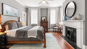 A bedroom with a bed, a ceiling fan, a fireplace, a mirror, and a painting on the wall. - Walk to Forsyth Park, Dining & Savannah Squares by Lucky Savannah (Savannah)