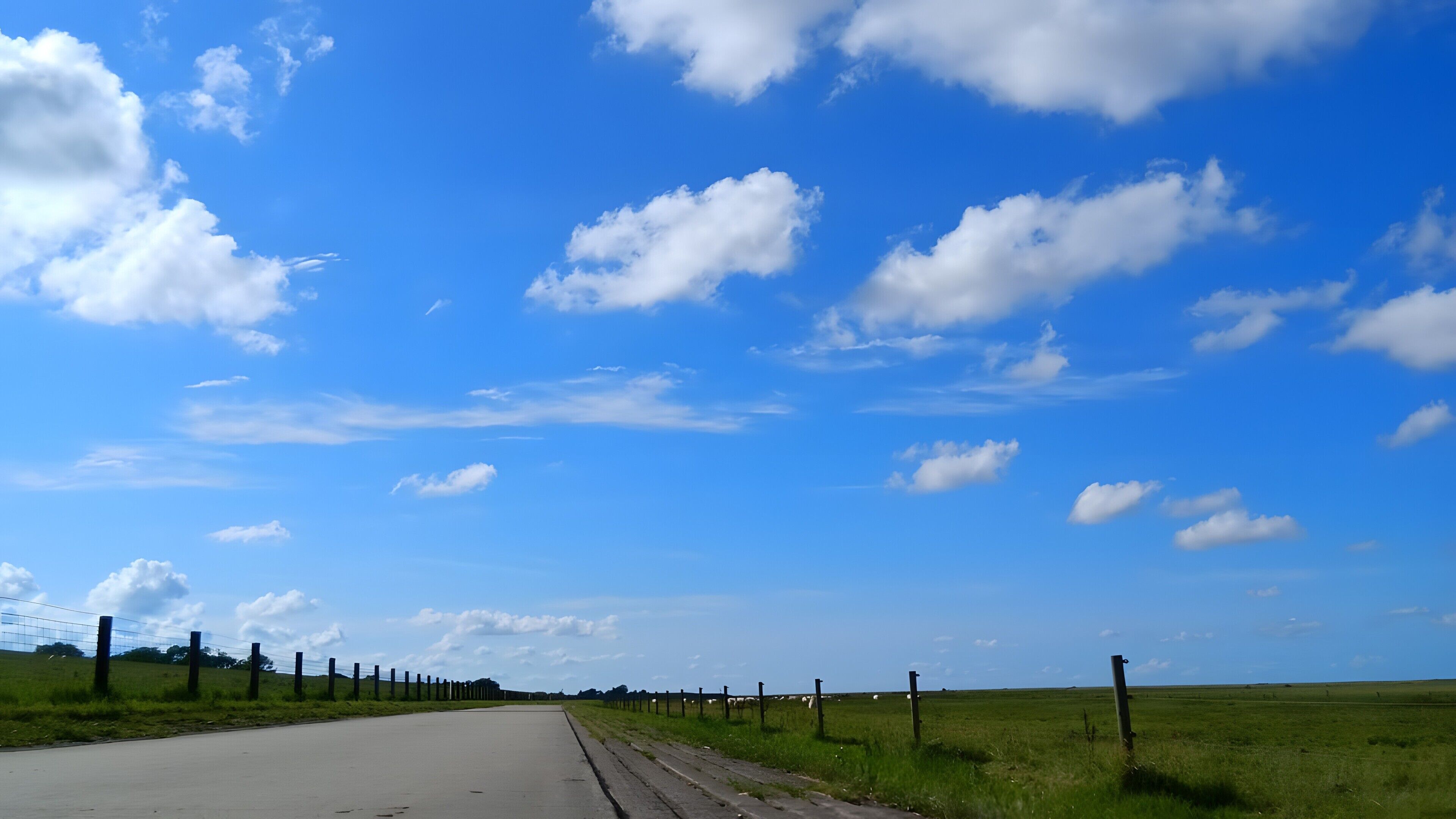 Une route de campagne, bordée d’un côté par une clôture, et un ciel bleu dégagé parsemé de nuages.