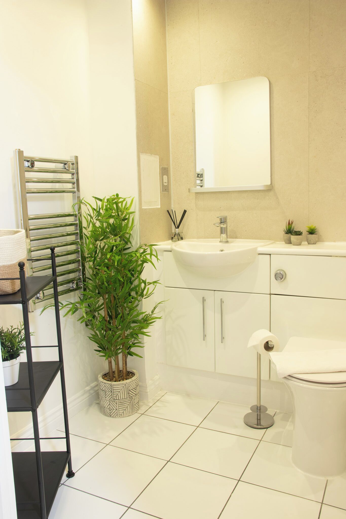 A modern bathroom with a white sink, mirror, and toilet. There is a towel rack, a potted plant, and a shelf with toiletries.