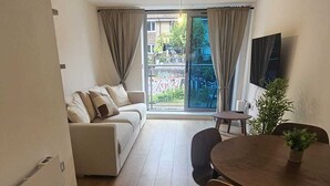 Ein modernes Wohnzimmer mit einer Couch, einem Esstisch mit Stühlen, einem Fernseher und einem großen Fenster mit Vorhängen.