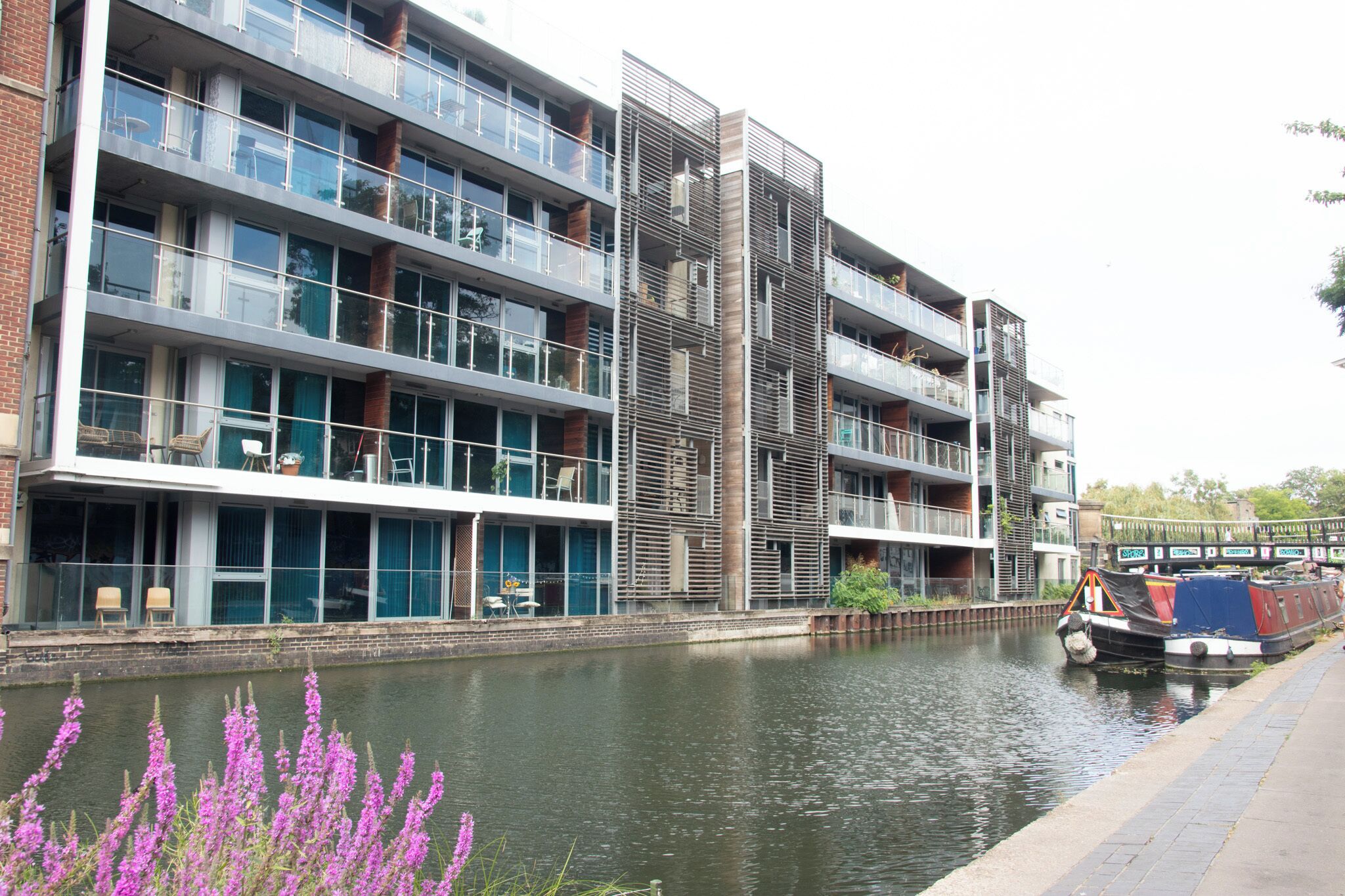 A modern multi-story building with balconies, a canal, and a narrowboat docked beside it.