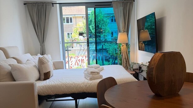 Ein modernes Hotelzimmer mit Bett, Sofa, Esstisch, Stühlen, einem großen Fenster mit Aussicht und einer Pendelleuchte.