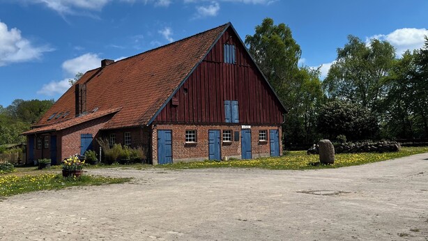 Une grange rouge avec un toit en pente, une allée en terre et un ciel bleu clair parsemé de nuages.