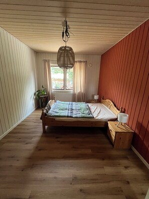 A bedroom with a wooden bed, a bedside table, a hanging light fixture, a window with curtains, and a potted plant. - Vacation at an Organic Farm (Schashagen)