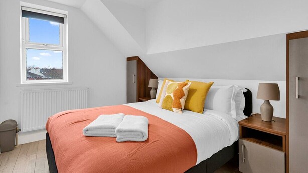 Un dormitorio con cama, almohadas, mesita de noche, lámpara, ventana y radiador.