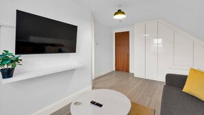 Una sala moderna con una televisión de pantalla plana, una mesa de centro redonda, un sofá con un cojín amarillo, una planta en maceta y una escalera que lleva a otra habitación.
