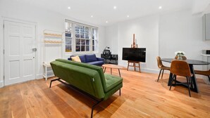 Un salon moderne avec un canapé vert, un divan bleu, une table à manger avec des chaises, un téléviseur à écran plat et un escalier visible à travers une grande fenêtre.