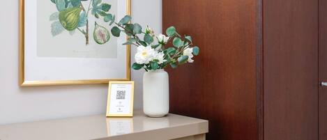 A framed botanical illustration, a white vase with flowers, and a framed welcome sign on a counter.