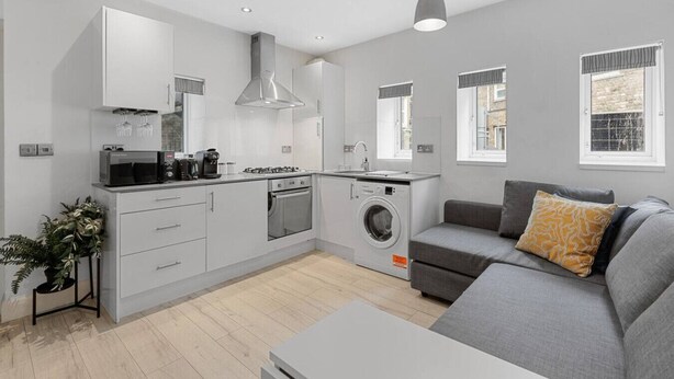 Eine moderne Küche mit Herd, Mikrowelle und Waschmaschine, ein graues Sofa mit gemustertem Kissen und eine kleine Pflanze.