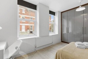 Une chambre avec une grande fenêtre, un radiateur, une armoire et un lit sur lequel est posée une serviette enroulée.
