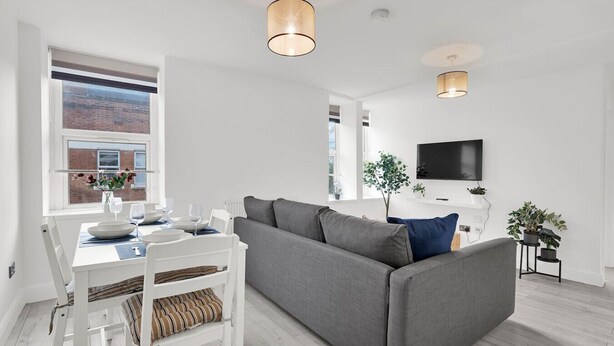 Ein modernes Wohnzimmer mit einem grauen Sofa, einem Esstisch mit weißen Stühlen und einem an der Wand befestigten Fernseher.