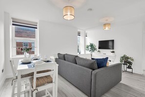 Ein modernes Wohnzimmer mit einem grauen Sofa, einem Esstisch mit weißen Stühlen und einem an der Wand befestigten Fernseher.