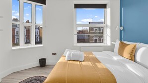 Ein Schlafzimmer mit einem großen Fenster, einem Bett mit weißer und goldener Bettwäsche und Blick auf Gebäude.