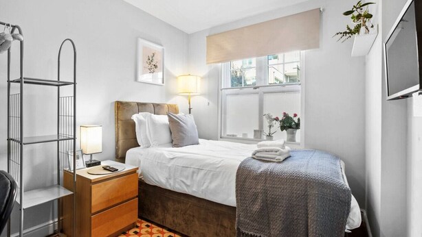 A bedroom with a bed, bedside table, lamp, and a window with a view. - Cosy Clapham Retreat - 2 Min Walk To Tube (Greater London)