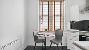 Una cocina moderna con una mesa de comedor, dos sillas, una ventana con persianas, horno empotrado y armarios blancos.