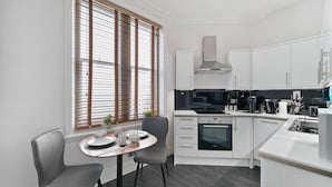 Una cocina moderna con muebles blancos, horno empotrado y comedor con mesa redonda y dos sillas.