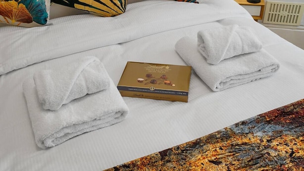 Una cama bien hecha con toallas dobladas, una caja de bombones y una alfombra estampada.