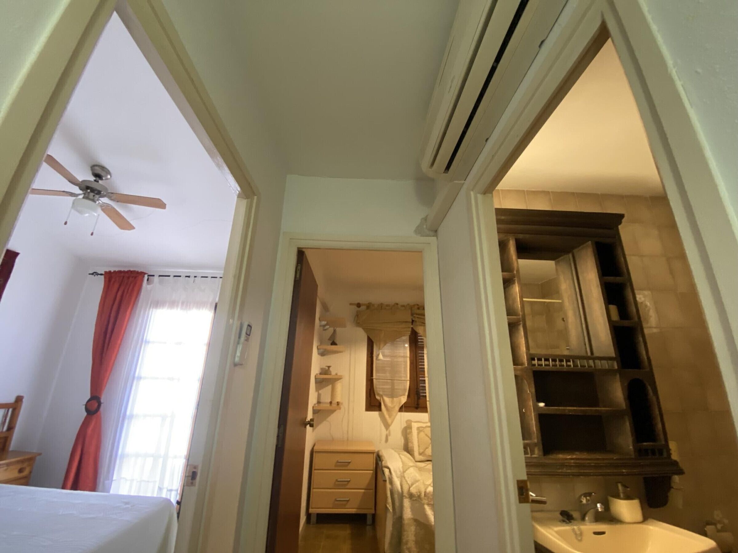 Un couloir étroit mène à une chambre à coucher équipée d’un lit, d’un ventilateur de plafond et d’une salle de bain avec un lavabo et un miroir.