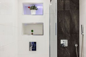 Ein Badezimmer mit weiß gefliesten Wänden, einem dunklen Duschbereich aus Marmor und drei Regalen mit Pflanzen und einem gerahmten Bild.