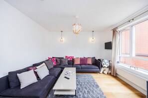 Ein modernes Wohnzimmer mit einem Ecksofa, einem Couchtisch, einem großen Fenster mit Vorhängen und einer dekorativen Hängelampe.