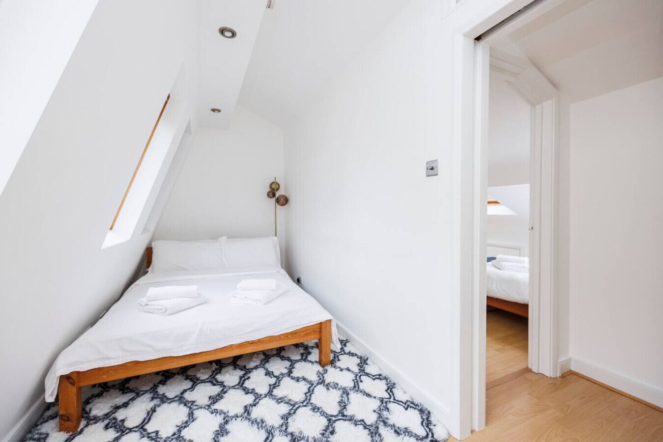 Ein kleiner, schräger Raum mit einem Bett, einem gemusterten Teppich und einer Tür zu einem anderen Zimmer.
