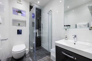 A modern bathroom with a glass-enclosed shower, a white toilet, a vanity with a mirror, and shelves with toiletries. - 3BR in Covent Garden Retreat - 6 people (Greater London)