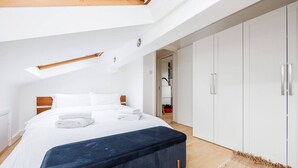 Un dormitorio moderno con tragaluz, una cama grande, un banco azul oscuro y armarios blancos.