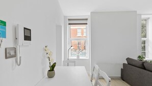 Ein moderner, minimalistischer Raum mit einem weißen Tisch, einem grauen Sofa, einer Topf-Orchidee, einem wandmontierten Telefon und einem Fenster mit Blick auf ein Backsteinhaus.