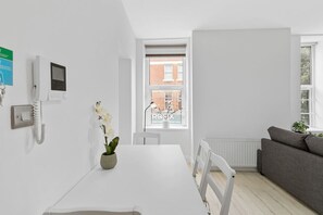 A modern, minimalistic room with a white table, a grey sofa, a potted orchid, a wall-mounted phone, and a window with a view of a brick building. - Perfectly Located 2BR Apartment in Balham (Greater London)