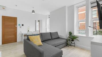 A modern living room with a grey sectional sofa, a wooden door, a dining area with a white table and chairs, and a large window with a view of a brick building.