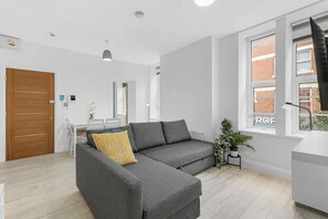 A modern living room with a grey sectional sofa, a wooden door, a dining area with a white table and chairs, and a large window with a view of a brick building. - Perfectly Located 2BR Apartment in Balham (Greater London)