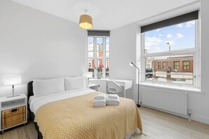A modern hotel room with a large bed, a desk, a chair, a radiator, and a window with a city view. - Perfectly Located 2BR Apartment in Balham (Greater London)