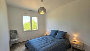 Un dormitorio con cama, dos mesitas de noche, una silla, un ventanal y una lámpara colgante.