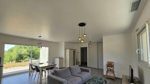 Amplia sala de estar con un sofá gris, una mesa de comedor con sillas y una cocina visible a través de una puerta abierta.