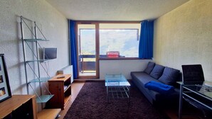 Un ambiente compatto con un divano, un tavolino, una TV fissata al muro e una finestra con vista sull'esterno.