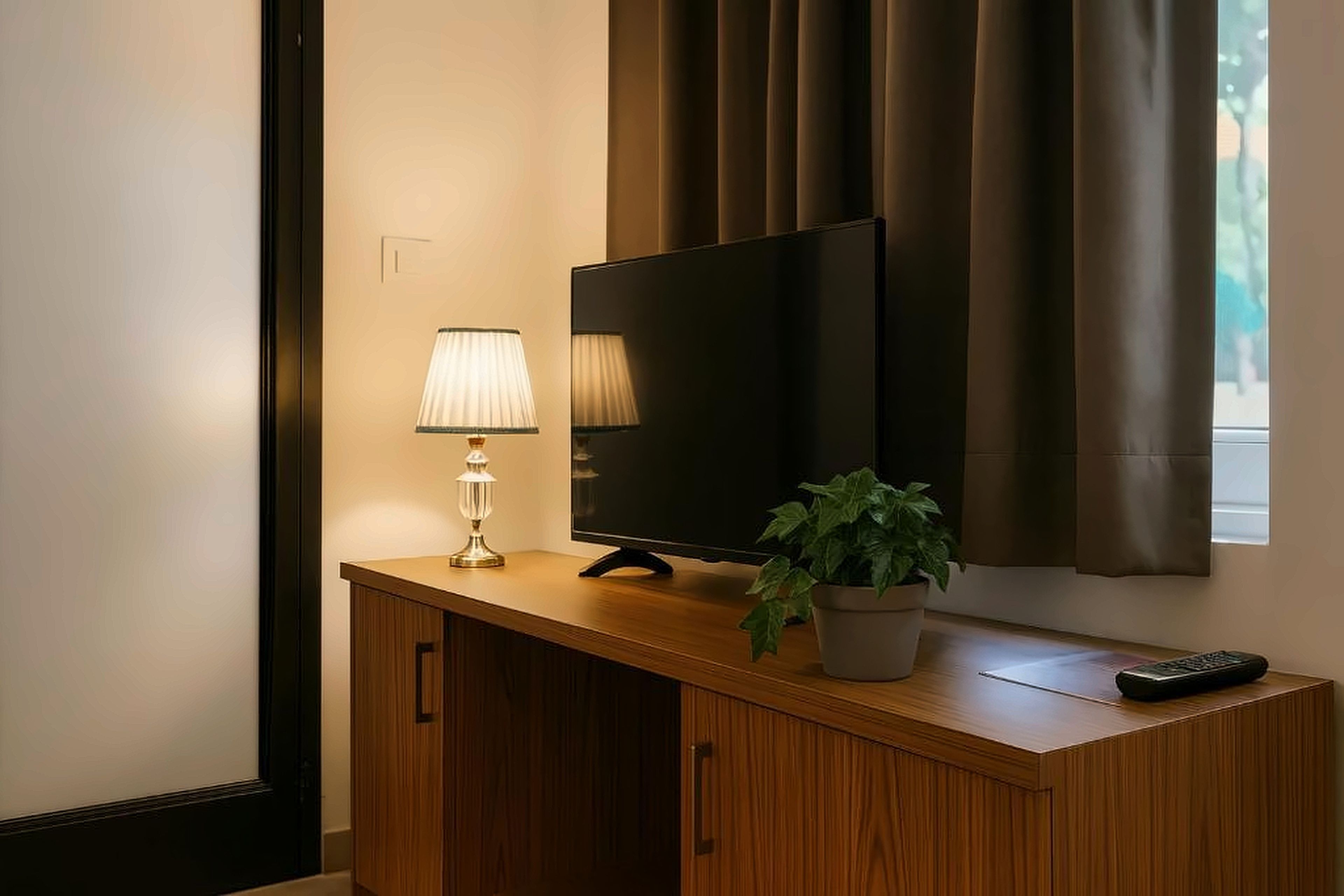 Una habitación de hotel con un escritorio de madera, una televisión, una lámpara, una planta en maceta y un control remoto.