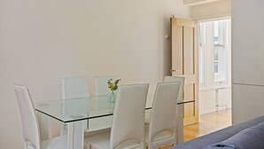 A modern dining area with a glass table and white chairs, a vase with yellow flowers, and a wooden door leading to a balcony. - 1BR Kensington | High St Kensington Tube&Hyde Park (Greater London)