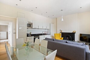 A modern kitchen with white cabinets, a microwave, and a dining area with a glass table and chairs. - 1BR Kensington | High St Kensington Tube&Hyde Park (Greater London)