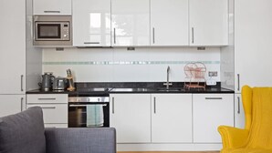 A compact kitchen with white cabinets, a microwave, a kettle, and a sink. There is a small oven and a chair with a yellow cushion. - 1BR Kensington | High St Kensington Tube&Hyde Park (Greater London)