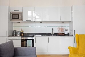 A compact kitchen with white cabinets, a microwave, a kettle, and a sink. There is a small oven and a chair with a yellow cushion. - 1BR Kensington | High St Kensington Tube&Hyde Park (Greater London)