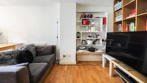 Un salón con un sofá gris, un mueble de televisión con televisor de pantalla plana, una estantería blanca con utensilios de cocina y suelo de madera.