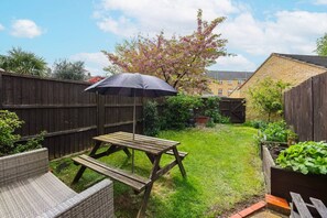 A backyard with a wooden table and bench, a picnic umbrella, and a garden bed with plants. - Double Ensuite 2 Min To Tube (Greater London)